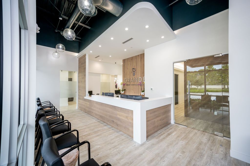 Open office layout view showing reception desk, waiting area with modern chairs, exposed ceiling elements, light wood flooring, and bright natural lighting