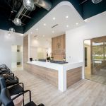 Open office layout view showing reception desk, waiting area with modern chairs, exposed ceiling elements, light wood flooring, and bright natural lighting