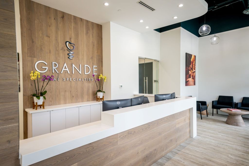 Reception area showing wood feature wall with GRANDE logo and heart symbol, white counter with floral arrangements, and contemporary waiting area seating