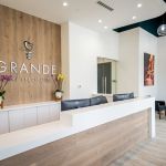 Reception area showing wood feature wall with GRANDE logo and heart symbol, white counter with floral arrangements, and contemporary waiting area seating