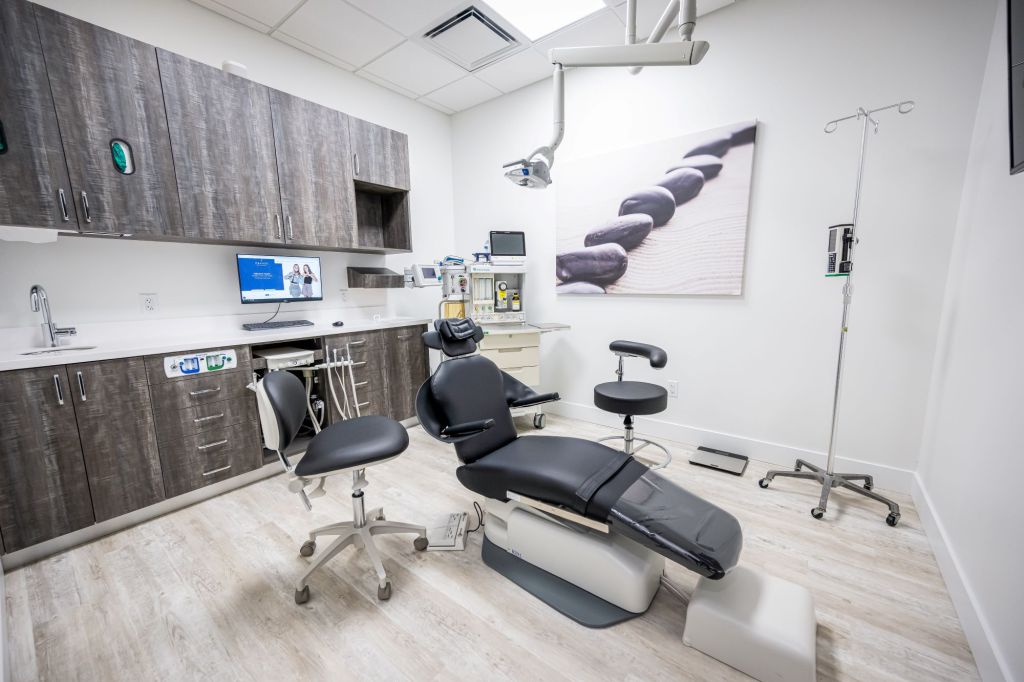 Dental treatment room with dark wood cabinetry, black patient chair, computer workstation, ceiling-mounted dental equipment, and light flooring