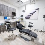 Dental treatment room with dark wood cabinetry, black patient chair, computer workstation, ceiling-mounted dental equipment, and light flooring